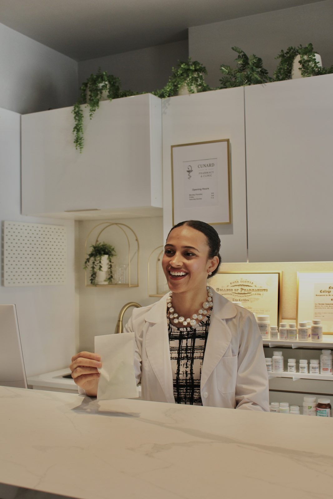 Cunard pharmacist handing a prescription bag across the dispensary counter, College of Pharmacists certificates behind